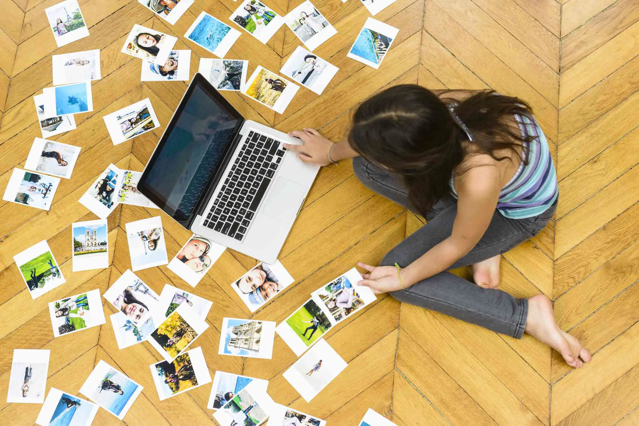 Alguém sentado no chão olhando fotos em um laptop com fotos espalhadas.