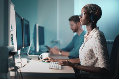 Closeup vista lateral de uma mulher negra loira de meados dos anos 20 fazendo seu projeto de desenvolvimento de software.  Ela está sentada na frente de um computador desktop e tomando um café enquanto trabalha em um computador.
