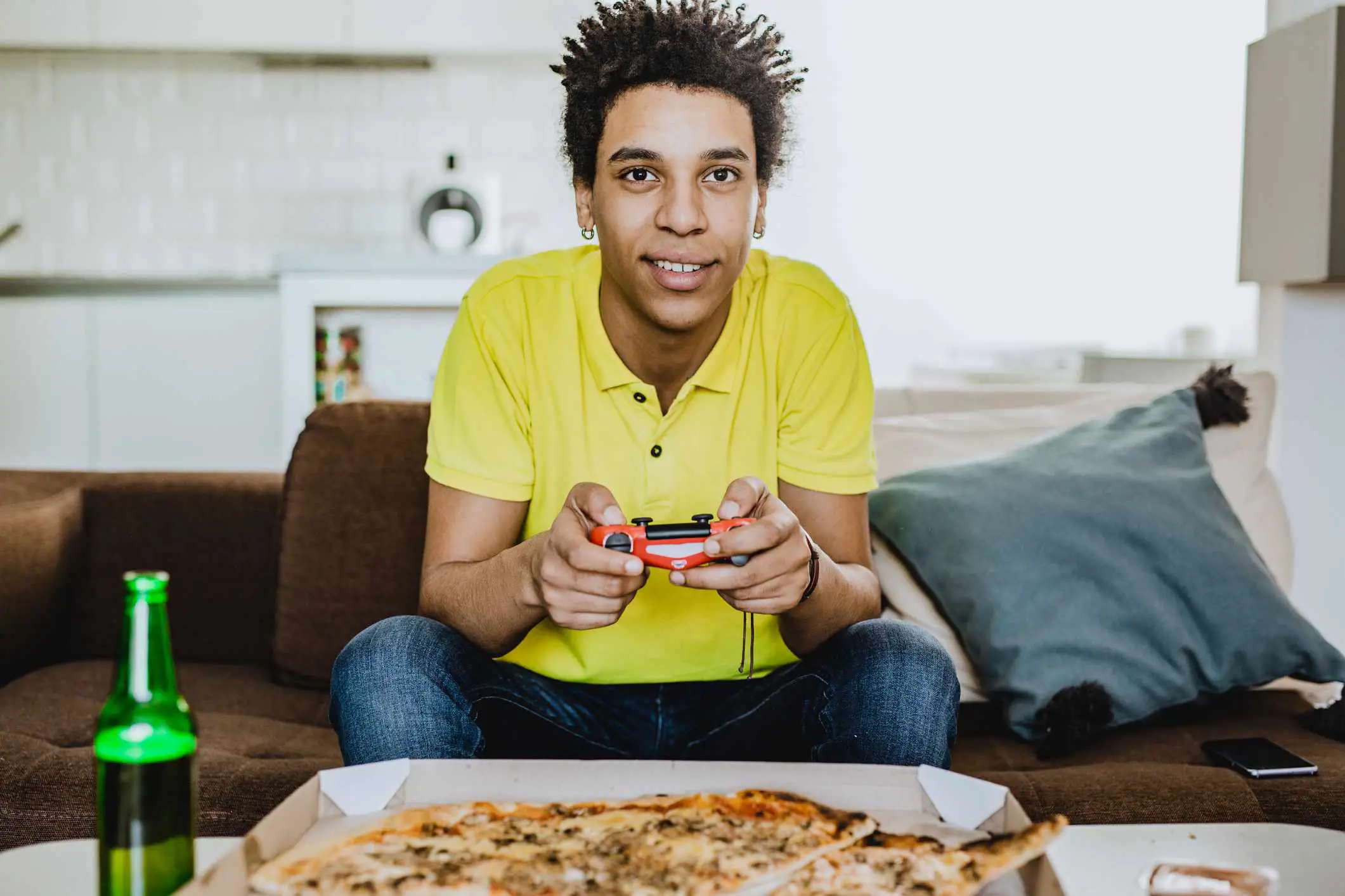 Um jovem adulto jogando videogame com uma pizza e uma bebida.