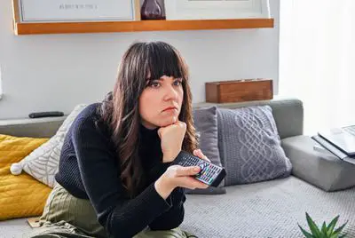 Uma mulher sentada, parecendo entediada com uma TV fora da tela, enquanto segura um controle remoto