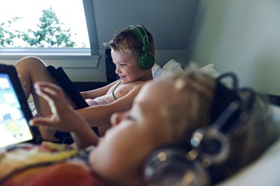 Duas crianças deitadas em uma cama com dispositivos de jogos nas mãos e fones de ouvido