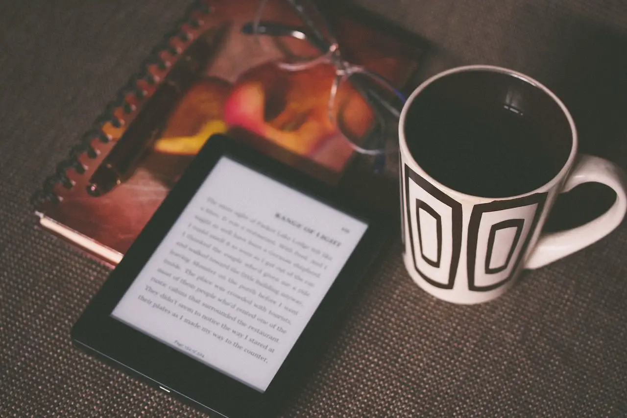 Imagem de um e-book, uma bebida, um caderno e um par de óculos em uma mesa