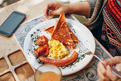 Perto de uma pessoa comendo um prato de café da manhã: torradas, ovos, tomates e salsicha em um prato.  Sobre a mesa, ao lado do prato, está um smartphone virado para cima e desligado.