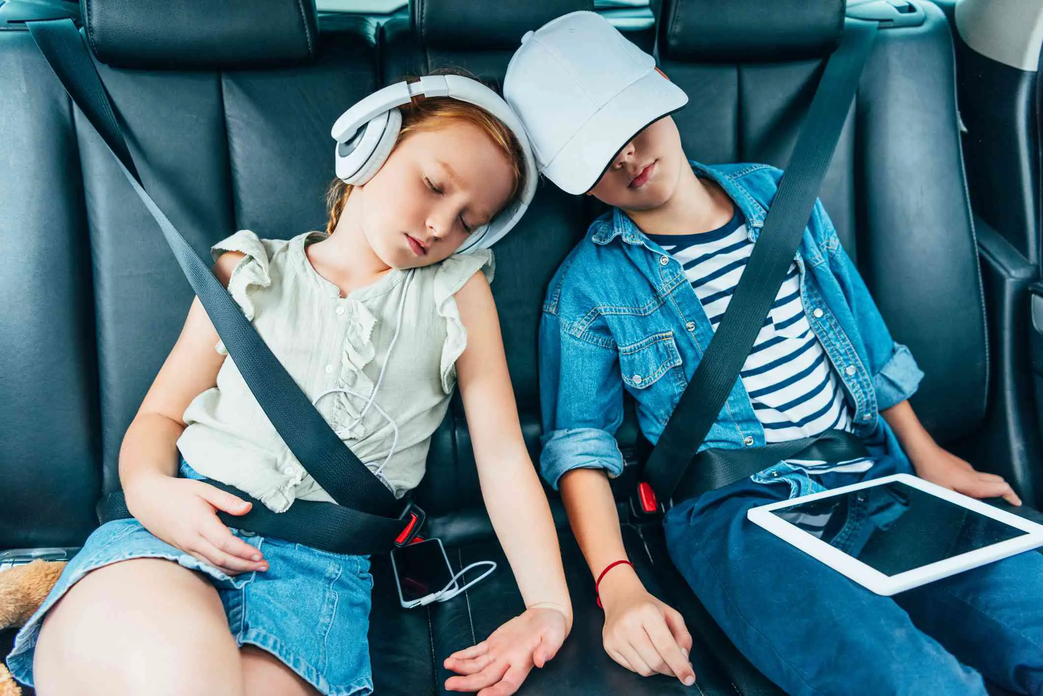 Duas crianças dormindo no carro segurando aparelhos eletrônicos que poderiam ser usados ​​para trabalhos escolares. 