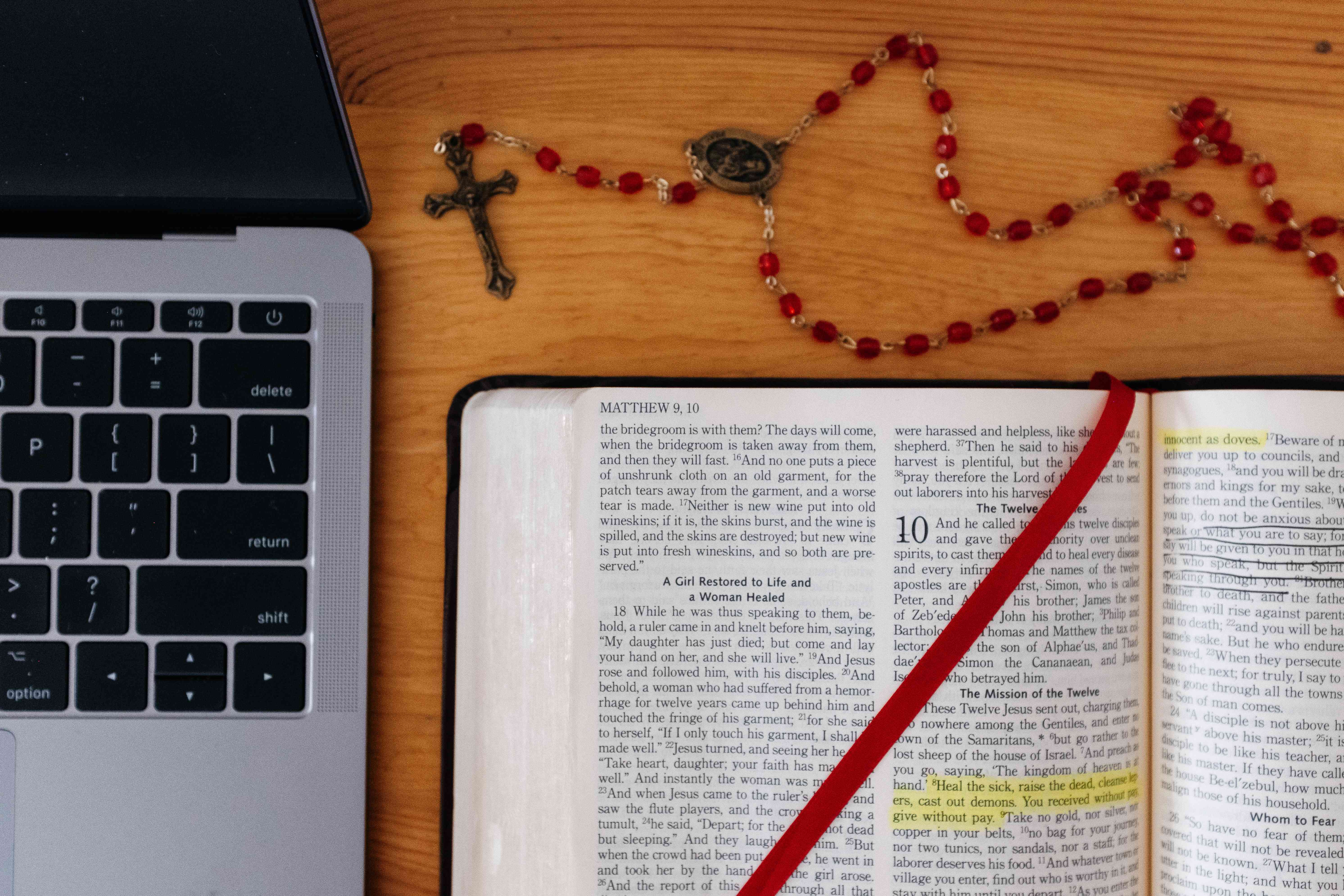 Uma Bíblia aberta e um rosário em uma mesa ao lado de um laptop. 