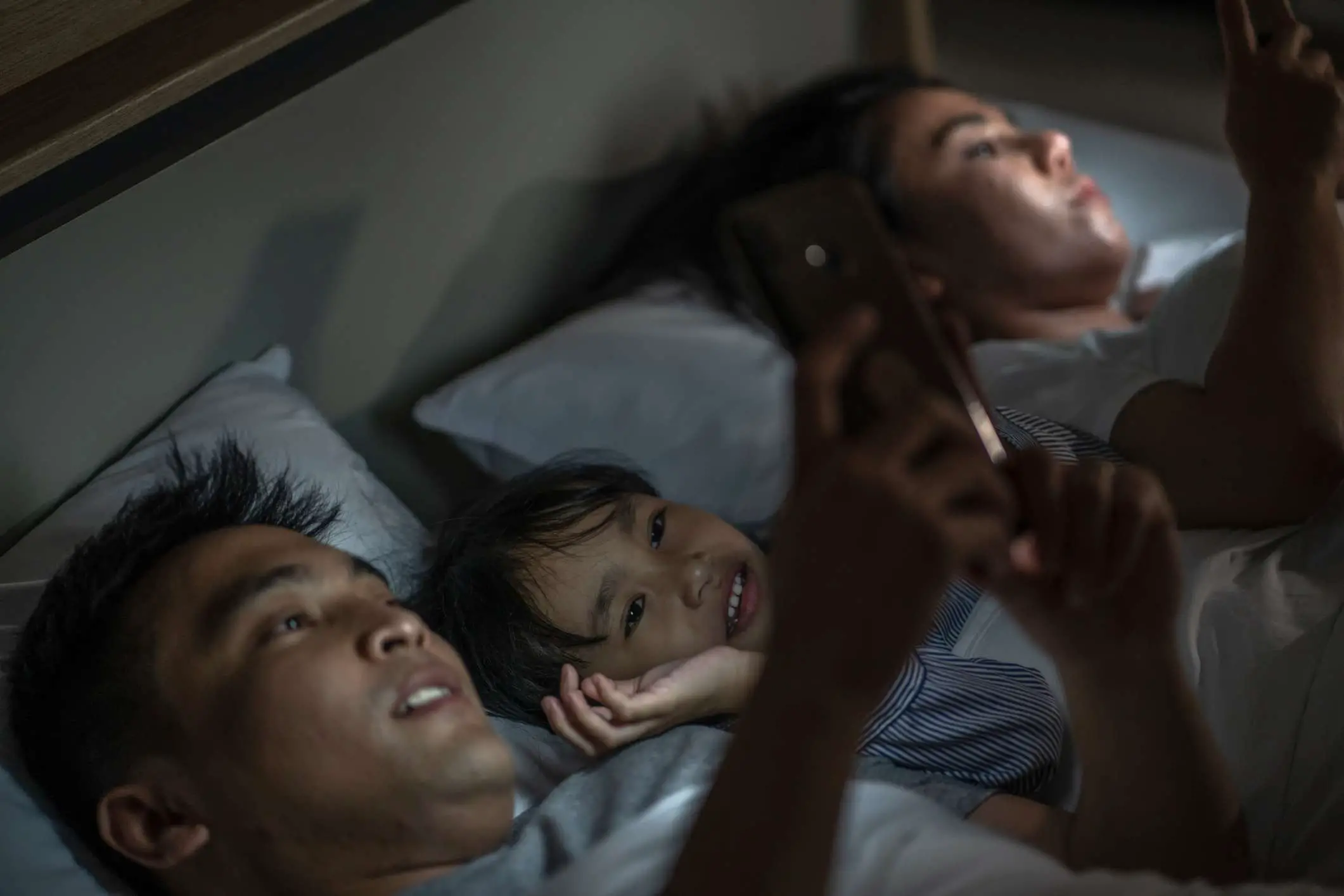 Uma família na cama com os pais brincando em smartphones e uma criança assistindo.