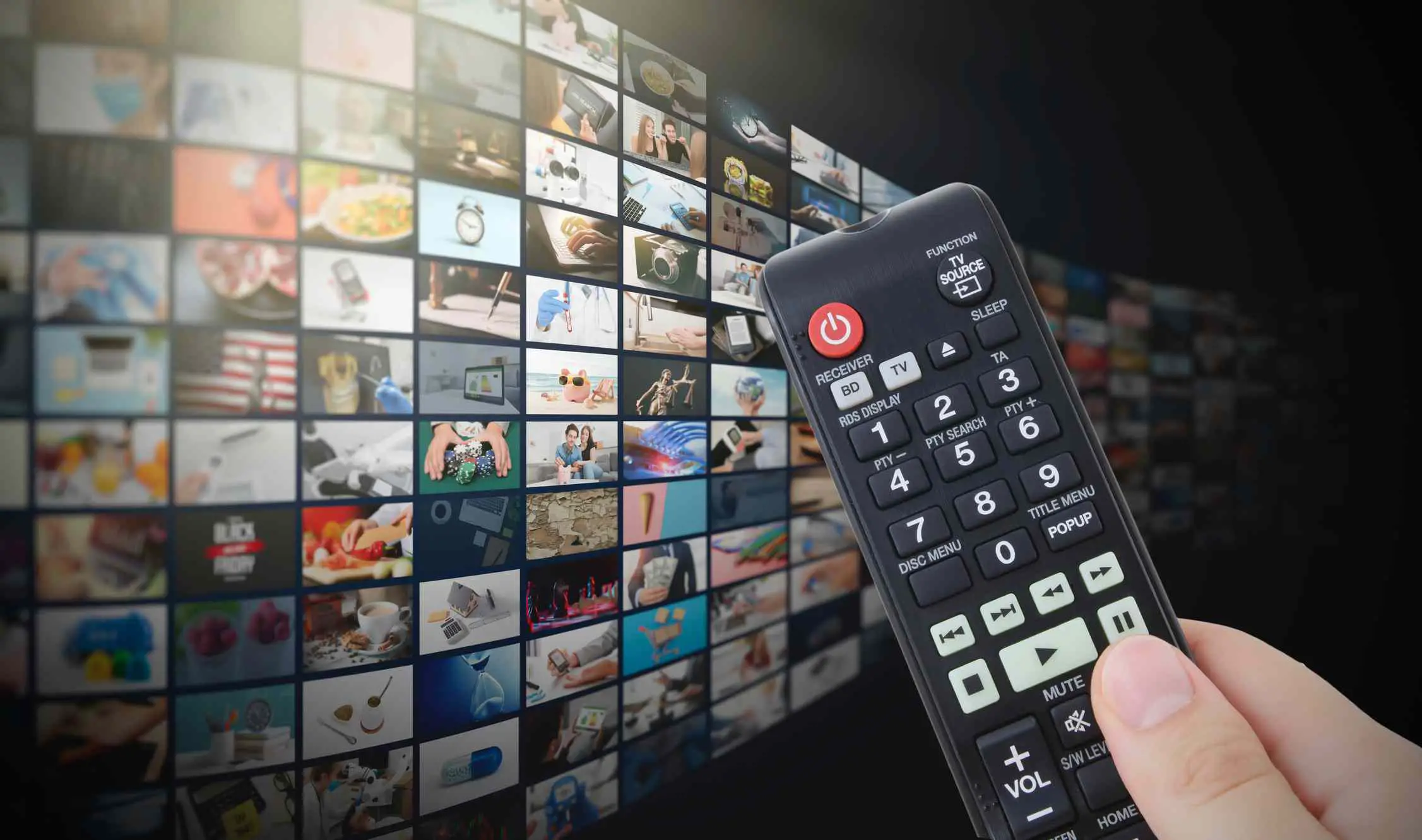 Uma mão segurando um controle remoto em frente a uma parede multimídia mostrando dezenas de canais de streaming.