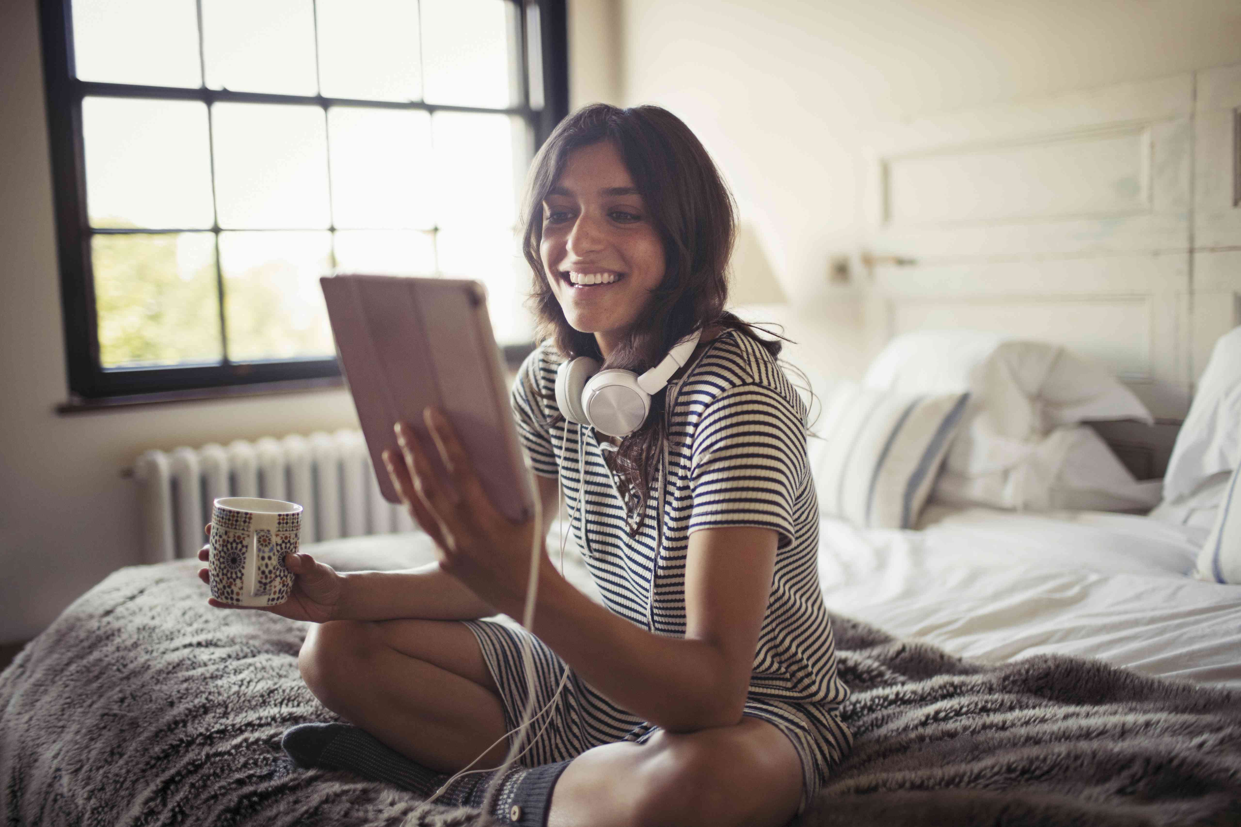 Mulher jovem sorridente com fones de ouvido, bebendo café e usando tablet digital na cama